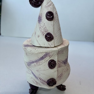 May include: A quirky ceramic ornament in cream with purple detailing. It has a conical hat with a swirl, three rose-shaped buttons, and a cylindrical base with three feet. The surface is textured with a leaf-like pattern.