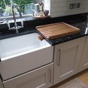 Puede incluir: Un fregadero blanco de estilo r&uacute;stico con un grifo cromado est&aacute; empotrado en una encimera de granito negro. Una tabla de secado de madera se encuentra en la encimera. La cocina tiene armarios blancos y una ventana con vegetaci&oacute;n.