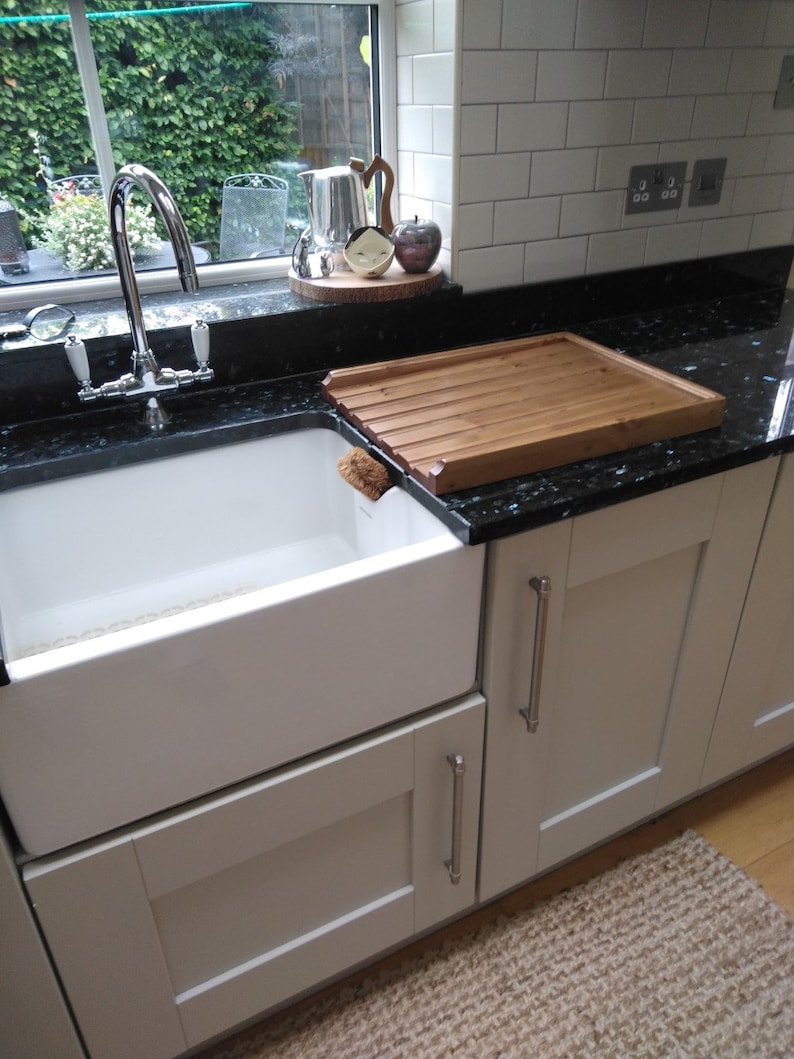 Puede incluir: Un fregadero blanco de estilo r&uacute;stico con un grifo cromado est&aacute; empotrado en una encimera de granito negro. Una tabla de secado de madera se encuentra en la encimera. La cocina tiene armarios blancos y una ventana con vegetaci&oacute;n.