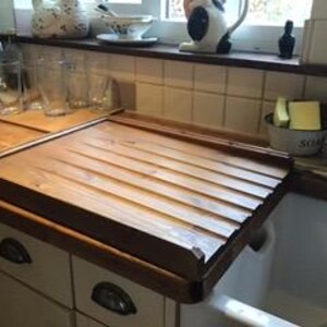 Puede incluir: Un escurreplatos rectangular de madera con dise&ntilde;o de listones, apoyado sobre un fregadero blanco. El estante es de madera marr&oacute;n claro y tiene un borde elevado. El entorno de la cocina incluye armarios, cristaler&iacute;a y productos de limpieza.