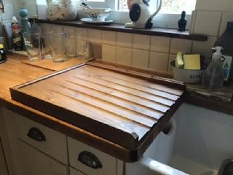 Puede incluir: Un escurreplatos rectangular de madera con dise&ntilde;o de listones, apoyado sobre un fregadero blanco. El estante es de madera marr&oacute;n claro y tiene un borde elevado. El entorno de la cocina incluye armarios, cristaler&iacute;a y productos de limpieza.