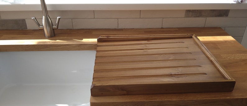 Puede incluir: Tabla de pan rectangular de madera clara con ranuras para recoger las migas. La tabla est&aacute; sobre una encimera de madera junto a un fregadero blanco. La tabla mide aproximadamente 38 cm por 30 cm.