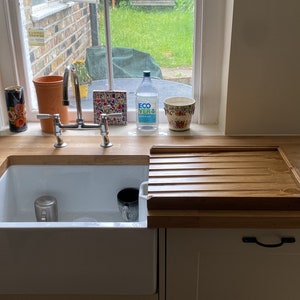 Puede incluir: Una zona de fregadero de cocina con un fregadero de porcelana blanca, encimera de madera y un escurreplatos de madera. Una ventana en el fondo con vistas a vegetaci&oacute;n. Varios objetos en el alf&eacute;izar, incluyendo una botella de agua y una taza floral.