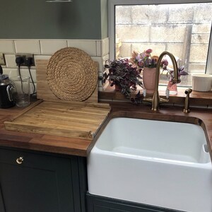 Puede incluir: Interior de cocina con fregadero blanco, encimeras de madera y armarios verde oscuro. Un salvamanteles tejido y una tabla de cortar de madera est&aacute;n sobre la encimera. Tambi&eacute;n se ve un grifo dorado y una l&aacute;mpara de pared verde.