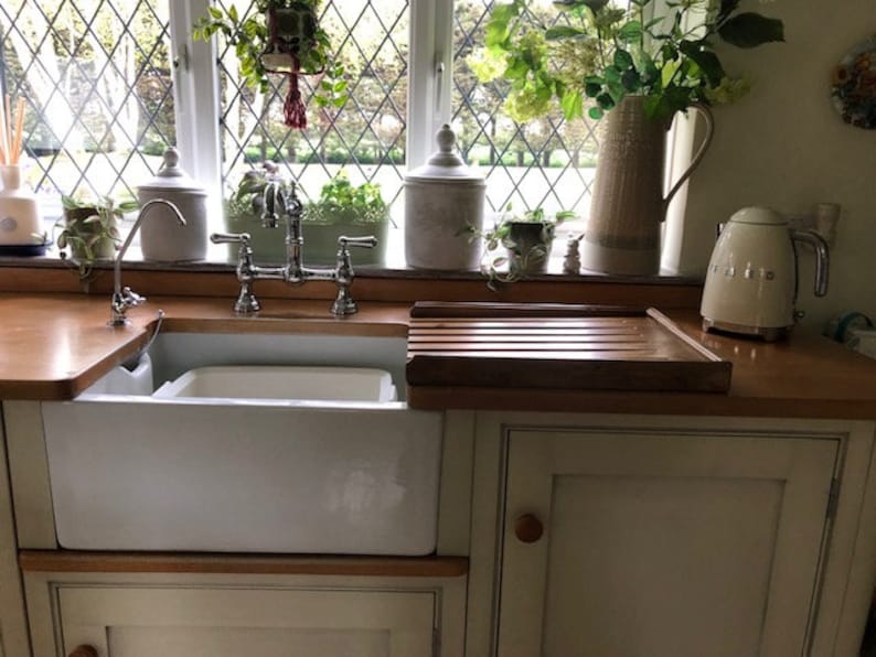 Puede incluir: Una cocina de estilo r&uacute;stico con un fregadero de porcelana blanca y un grifo cromado. Una tabla de cortar de madera y un hervidor el&eacute;ctrico blanco se encuentran sobre la encimera de madera. Plantas y flores verdes adornan la ventana.