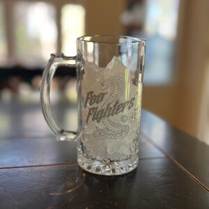 May include: A clear glass beer stein with a handle, filled with ice cubes. The stein has a graphic of a snake and the text "Foo Fighters" etched on the side.