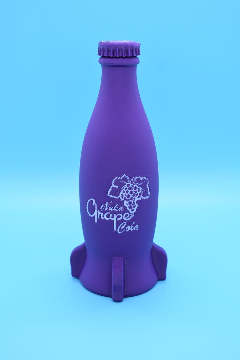 May include: A purple glass bottle shaped like a rocket with a white label that says "Nuka Grape Cola" and a picture of a bunch of grapes.
