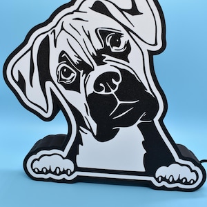 May include: Black and white silhouette of a boxer dog peeking over a black base. The dog's head is tilted to the side, and its eyes are looking directly at the viewer.