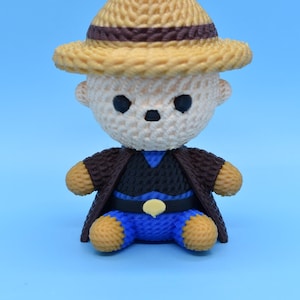 May include: A handcrafted, crocheted bear figurine wearing a brown hat, jacket, and blue pants. The bear has black eyes and a small nose. The hat and bear's limbs are tan, while the jacket is dark brown. The bear is sitting on a light blue surface.