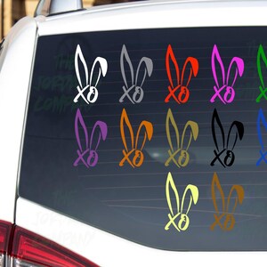 May include: A variety of colorful bunny ear and "XO" stickers are displayed on a car window. The stickers are in white, gray, red, pink, green, purple, orange, gold, black, yellow, and blue. The stickers are likely for Easter or spring.