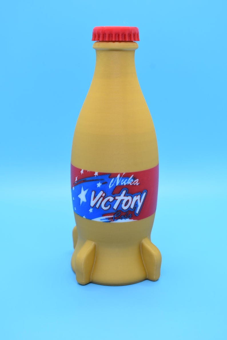May include: A yellow Nuka-Cola Victory bottle with a red cap and a rocket-shaped base. The bottle has a red, white, and blue label with the words "Nuka-Cola Victory" printed on it.