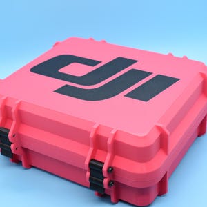 May include: A bright pink hard-shell case with a black DJI logo. The case has a latch on the side and is made of durable plastic.