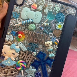 May include: A navy blue tablet case decorated with numerous charms, including a large blue cross, a Hello Kitty figure, bows, and a rainbow lollipop. The case features the text "PRETTY LADYR" and "BABY".