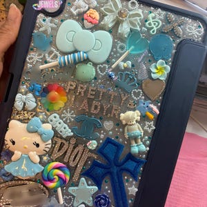 May include: A dark blue tablet case decorated with a variety of light blue and multicoloured charms, including a Hello Kitty figure, a large bow, a blue cross, lollipops, and text that reads PRETTY LADYR, BABY, and DIOR, all set on a glittery base.