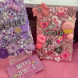 Può includere: Due custodie per telefono decorate, una viola e una rosa, ricoperte di perle, ciondoli ed elementi decorativi. La custodia viola ha la scritta "EMNEW" e un lecca-lecca. La custodia rosa ha la scritta "LADYR" e un cupcake.