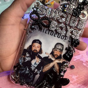 Può includere: Custodia trasparente per telefono con la scritta "SUICIDE BOYS" in strass argentati. La custodia presenta un'immagine in bianco e nero di due uomini, completata da elementi decorativi neri e un ciondolo a forma di spada argentata.