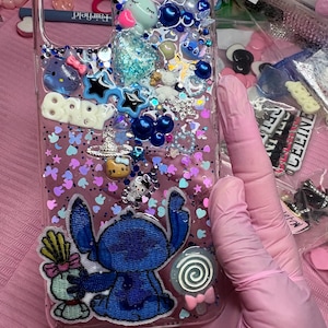 Puede incluir: Funda de teléfono transparente decorada con adornos azules, blancos y rosas. Presenta un diseño del personaje Stitch azul con una piruleta y un pequeño personaje. Incluye estrellas, cuentas y purpurina.