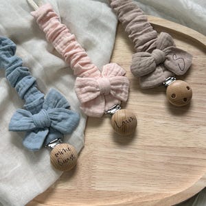 Könnte beinhalten: Drei Schnullerketten in Blau, Hellrosa und Beige, jeweils mit einem gerafften Stoffband, einer dekorativen Stoffschleife und einem runden Holzclip. Die Holzclips sind mit dem Text mini Quast, Lavin und dem Buchstaben S graviert.