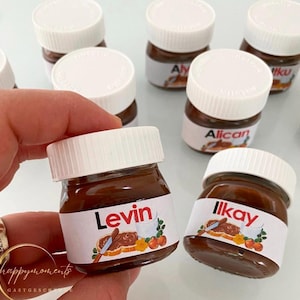 Könnte beinhalten: Eine Sammlung kleiner Glasgläser gefüllt mit Schokoladenaufstrich, jedes mit einem personalisierten Etikett mit einem Namen. Die Gläser haben weiße Deckel und sind auf einer weißen Oberfläche angeordnet.