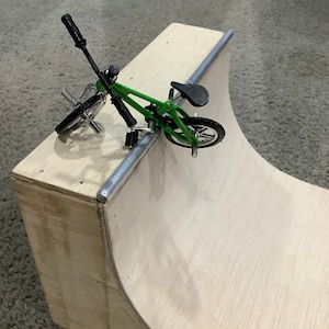 May include: A green toy BMX bike with black handlebars and wheels is positioned on a wooden ramp. The ramp is made of plywood and has a curved edge.