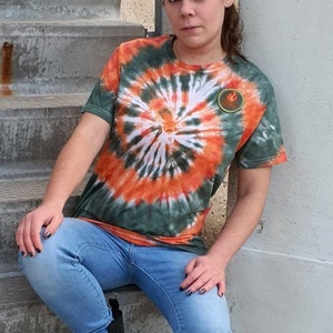 Op de afbeelding: Een vrouw draagt een groen, oranje en wit tie-dye T-shirt met een klein rond ontwerp aan de voorkant. Ze draagt een blauwe jeans en zwarte combat boots. Ze zit op een betonnen trap.