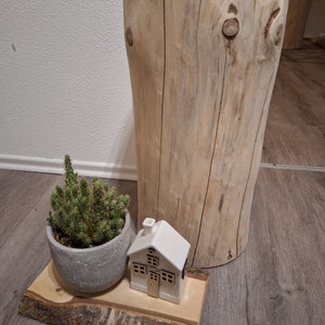Könnte beinhalten: Ein Baumstumpf aus Holz mit natürlicher Rindenoptik, der als Hocker verwendet wird. Eine kleine Keramik-Hausfigur und eine Topfpflanze mit Sukkulenten stehen auf einer Holzscheibe vor dem Baumstumpf.