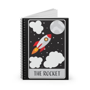 May include: Spiral-bound notebook with a black cover featuring a rocket ship design. The rocket is white and red with a Bitcoin symbol. The background is black with clouds, stars, and a moon. The text "THE ROCKET" is at the bottom.