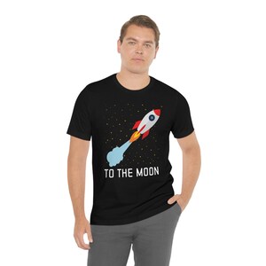 May include: Black t-shirt featuring a cartoon rocket blasting off with the text "TO THE MOON" in white. The rocket is white and red with yellow and orange flames, set against a starry background.