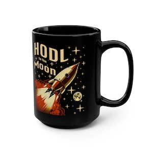 May include: Black ceramic mug featuring a retro rocket ship design. The mug has the text "HODL to the Moon" with stars and a moon. The rocket is illustrated with red and gold accents. The mug has a large handle.