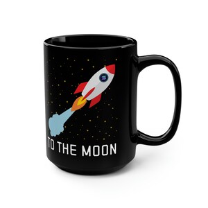 May include: Black ceramic mug featuring a rocket ship graphic with the text "TO THE MOON." The rocket is white and red, with a blue exhaust trail and a starry background. The mug has a large handle.