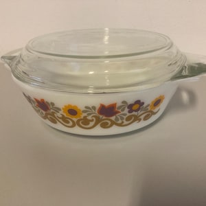 May include: A round white milk glass casserole dish with a clear glass lid and integrated side handles. The exterior features a decorative band of stylised orange, yellow, and purple flowers with brown scrolling foliage patterns around the base.