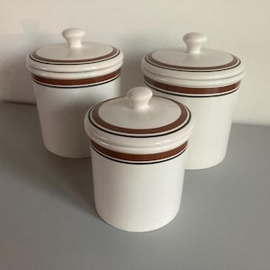 Op de afbeelding: Een set van drie witte keramische voorraadpotten met bijpassende deksels. Elke pot is voorzien van een horizontale bruine band met dunne zwarte lijnen die zowel het deksel als de bovenkant van de cilindrische behuizing omcirkelen.