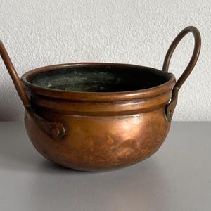 May include: A round, hammered copper bowl featuring two curved metal handles attached to the sides with rivets. The exterior has a warm, metallic copper finish, while the interior shows signs of oxidation with a dark, greenish-patina finish.