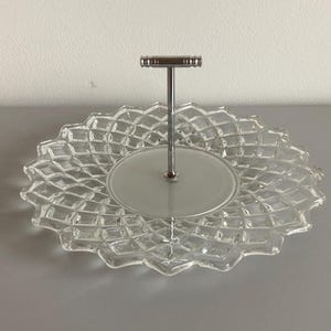 Op de afbeelding: Glazen serveerschaal met een ruitpatroon en een zilveren metalen handvat. De schaal heeft een ronde basis en een decoratieve, geschulpte rand. Het handvat is bevestigd in het midden van de schaal.
