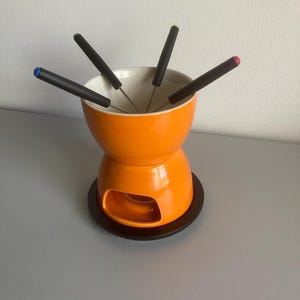 Op de afbeelding: Een oranje keramische fonduepan met een zwarte basis en zes fonduevorken. De vorken hebben zwarte handvatten met gekleurde uiteinden: blauw, rood en andere. De binnenkant van de pan is wit.