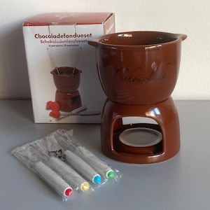Op de afbeelding: Een bruine keramische chocoladefondueset met kom en voet, vergezeld van vier fonduevorkjes met witte handvatten met rode, gele, blauwe en groene uiteinden, verpakt in een doos met de tekst Chocoladefondueset 4-persoons.