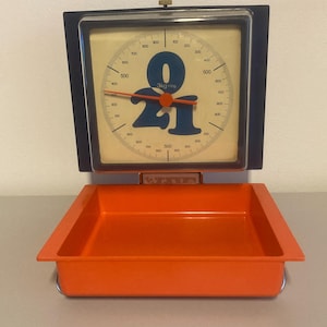 May include: Vintage orange and black kitchen scale with a square face and a dial displaying numbers and measurements up to 1000 grams. The dial features the number "021" in blue. The scale has a matching orange tray.