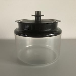 Könnte beinhalten: Ein klares Glasgefäß mit schwarzem Deckel und Griff. Das Gefäß hat eine abgerundete Form und eine weite Öffnung. Der Deckel ist flach mit einem kleinen, zylindrischen Griff oben. Das Gefäß wird wahrscheinlich zur Aufbewahrung verwendet.