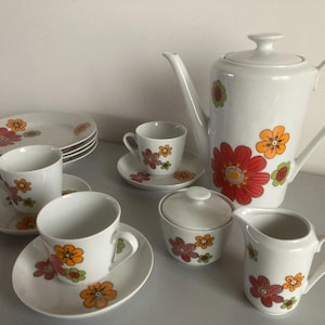Può includere: Servizio da tè in ceramica bianca con un motivo floreale arancione, rosso e verde. Il set include una teiera, tazze, piattini, piatti, una zuccheriera e una lattiera. Il motivo floreale è ripetuto.