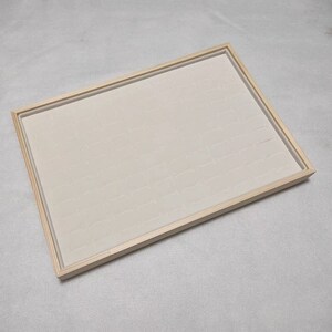 May include: A wooden ring display tray with a light brown frame and a white felt interior. The tray has many small compartments for storing rings.