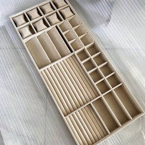 May include: A beige velvet jewelry organizer with compartments for rings, bracelets, necklaces, and watches. The organizer has a variety of compartments, including a ring roll, a watch pillow, and a necklace holder.
