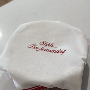 May include: A round, off-white fabric cover featuring the embroidered red text "Shhh... I'm fermenting" centered on the top surface. The fabric has a visible woven texture and is draped over a circular object, with a red striped edge visible at the base.