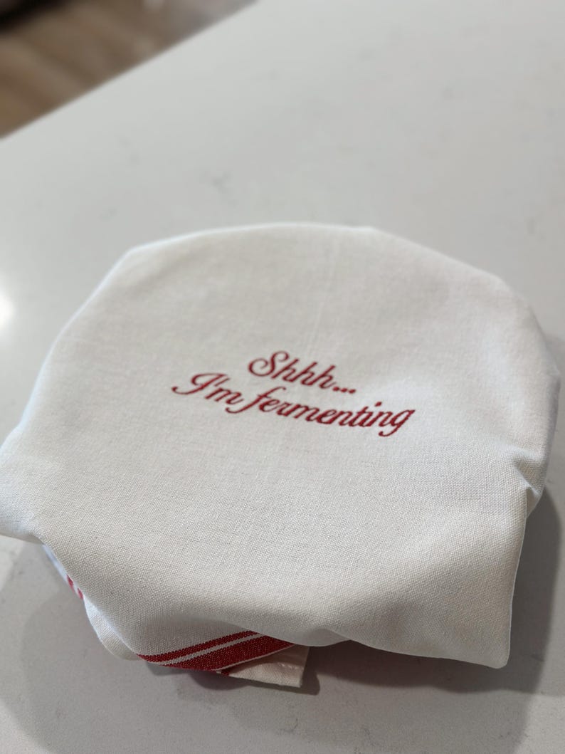 May include: A round, off-white fabric cover featuring the embroidered red text "Shhh... I'm fermenting" centered on the top surface. The fabric has a visible woven texture and is draped over a circular object, with a red striped edge visible at the base.