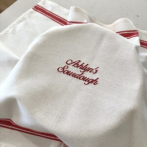 Puede incluir: Pa&ntilde;o de cocina de lino blanco con rayas rojas y texto bordado "Ashlyn's Sourdough". La toalla est&aacute; doblada, con el texto bordado visible. Las rayas rojas est&aacute;n a lo largo de los bordes.