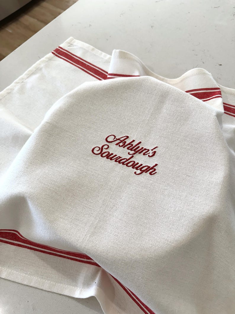 Puede incluir: Pa&ntilde;o de cocina de lino blanco con rayas rojas y texto bordado "Ashlyn's Sourdough". La toalla est&aacute; doblada, con el texto bordado visible. Las rayas rojas est&aacute;n a lo largo de los bordes.