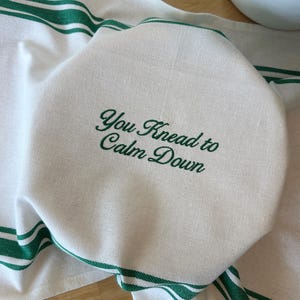 Puede incluir: Paño de cocina blanco con rayas verdes y la frase bordada "You Knead to Calm Down" en verde. El paño está doblado sobre un objeto redondo, posiblemente masa, sobre una superficie de madera.