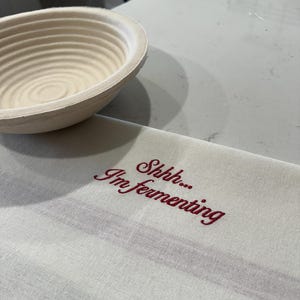 May include: A round, beige, ribbed proofing basket made of natural cane, positioned next to a rectangular, off-white kitchen towel featuring the embroidered red text "Shhh... I'm fermenting" in a cursive script, with a subtle horizontal stripe.