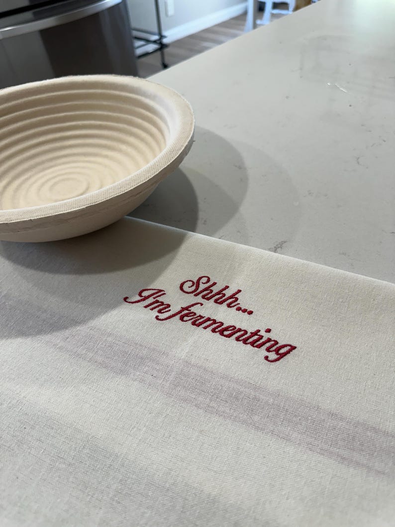 May include: A round, beige, ribbed proofing basket made of natural cane, positioned next to a rectangular, off-white kitchen towel featuring the embroidered red text "Shhh... I'm fermenting" in a cursive script, with a subtle horizontal stripe.