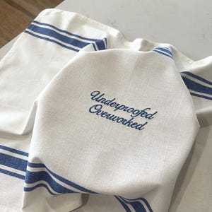 May include: White kitchen towel with blue stripes and the embroidered text "Underproofed Overworked". The towel is folded and lies on a light-colored surface. The design is simple and classic.