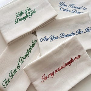 May include: Five off-white rectangular kitchen towels featuring embroidered text. The towels display the phrases Life of a Dough Girl in green, You Knead to Calm Down in blue, Are You Bready for It? in blue, The Fate of Doughphelia in green, and In my sourdough era in red.